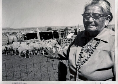 Black and white photograph of Dorthea Begay, an older woman in glasses wearing a large round cluster brooch and a long silver necklace. She stands beside a wire fence with a flock of sheep in a corral behind her and open desert in the distance.