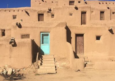 Multistory adobe building at Taos Pueblo under a deep blue sky, with small wooden windows, a turquoise door at the foot of a short staircase, and dry earth in the foreground.