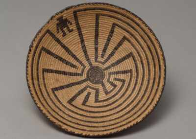 Round coiled basketry tray in natural tan with a black woven design of the Man in the Maze: a small human figure stands at the entrance of a labyrinth that winds inward toward the center.