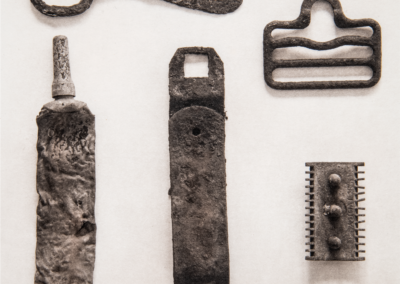 Platinum palladium print of six worn, rusted metal objects arranged on a pale background, including a bottle opener, a flattened tube, a buckle, a strap fragment, a razor head, and a narrow blade.