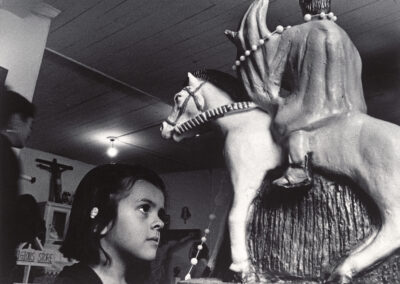 Black and white photograph of a young girl with dark hair looking up at a carved statue of Santiago on horseback, draped in strands of beads, inside a church in Chimayó.